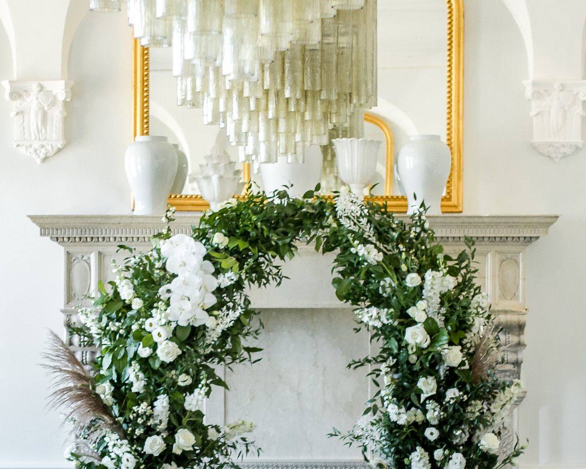 An opulent floral archway arrangement, highlighting the beatuful marble mantlepiece below a chandelier.