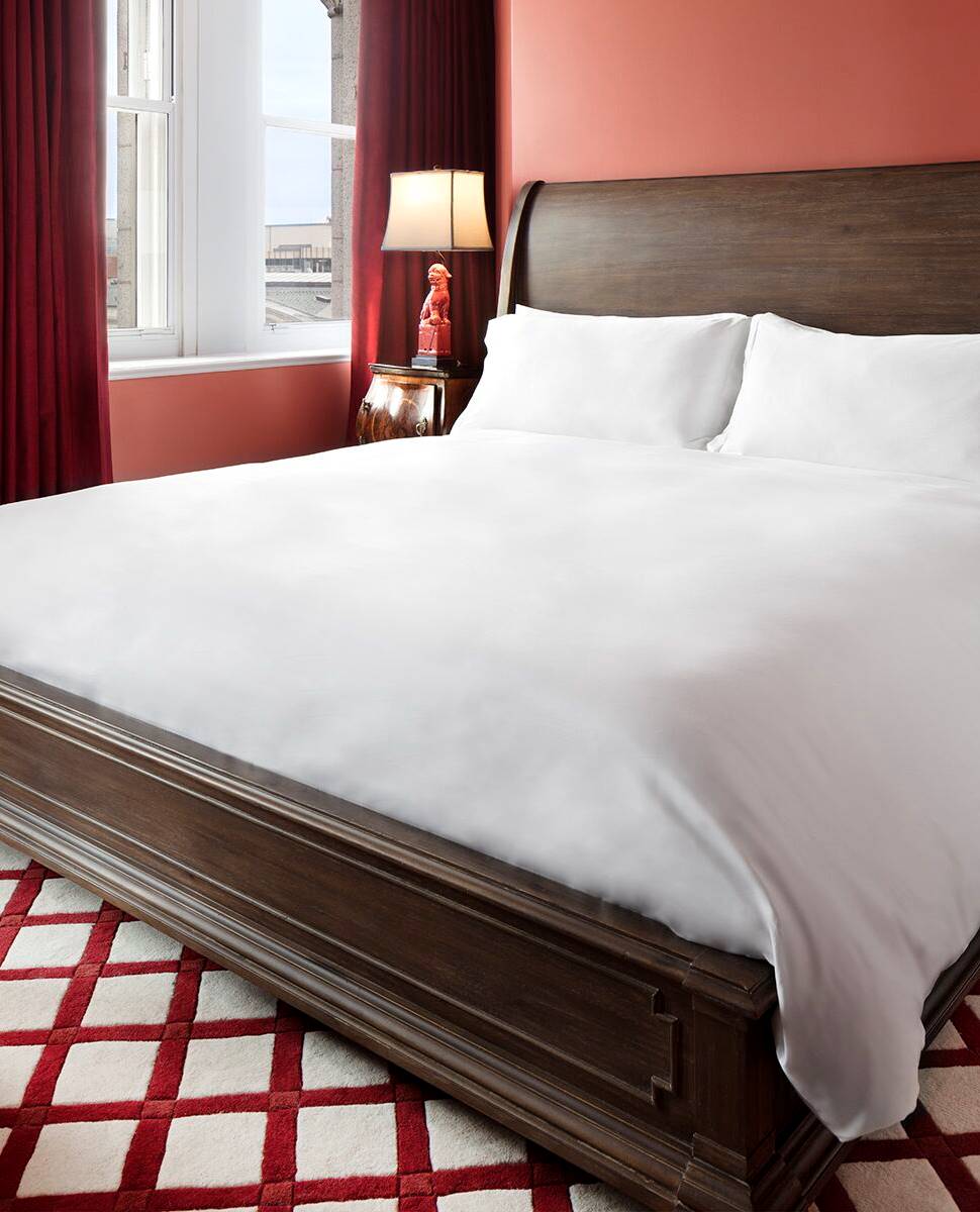 Bedroom with dark wood bed frame, white bedding, and red and white checkered rug near tall windows at Riggs DC.
