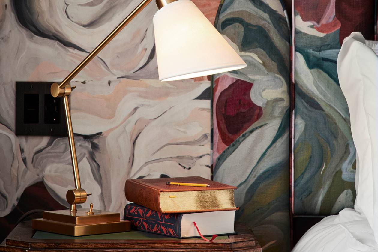 Bedside table at a Riggs DC hotel room with stacked books, a brass reading lamp, and abstract floral wallpaper backdrop.